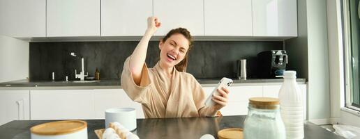 Attractive young cheerful girl baking at the kitchen, making dough, holding recipe book, having ideas photo