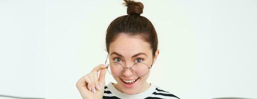 Image of woman in glasses, looks surprised, takes off eyewear, looks amazed, says wow, impressed by something, stands over white background photo