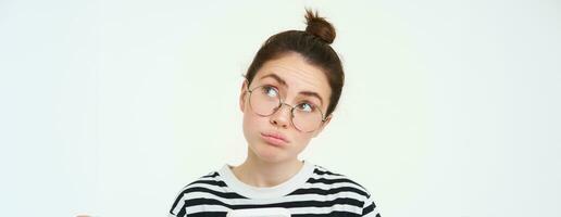 Close up portrait of girl in glasses, looks puzzled, holds phone and thinks, makes choice, decides what to order, does online shopping in mobile app, white background photo