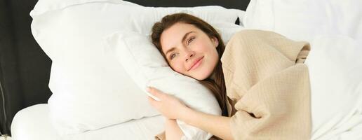 maravilloso mujer descansando en cama vistiendo pijama, abrazando su almohada y sonriente foto