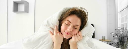 Portrait of girl feeling sleepy after waking up early, lying in bed under blanket and smiling photo