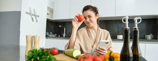 imagen de joven hermosa mujer, participación tomate, sentado en cocina con teléfono inteligente, el cortar tablero y vegetales en encimera, Cocinando alimento, orden comestibles para su receta, utilizando móvil teléfono aplicación foto