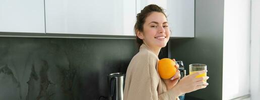 Beautiful young woman in bathrobe, drinking homemade orange juice, smiling and laughing, standing near worktop in kitchen photo