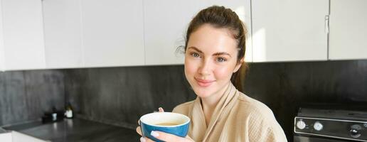 guapo joven mujer con taza de café, posando en el cocina, sonriente a cámara, disfruta su Mañana a hogar, vistiendo acogedor bata de baño foto