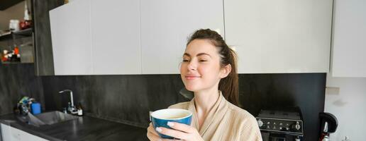 Portrait of smiling woman drinks coffee, stands in the kitchen and enjoys delicious cup of cappuccino in the morning, looks happy at camera photo