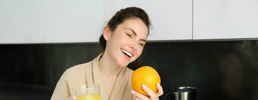 retrato de atractivo joven moderno mujer, posando en cocina, participación vaso de jugo y un naranja, riendo y hablando a alguien aparte foto