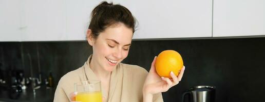 Portrait of beautiful, healthy young woman with orange in hand, drinks freshly squeezed juice from glass, posing in kitchen, enjoys her morning at home photo