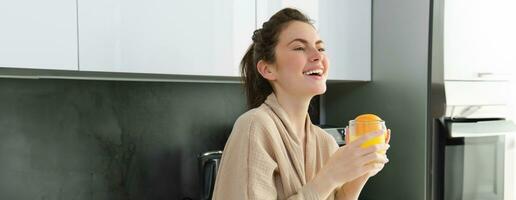 retrato de atractivo joven mujer riendo en cocina, vistiendo bata de baño, Bebiendo naranja jugo, hecho en casa beber, participación vaso y sonriente foto