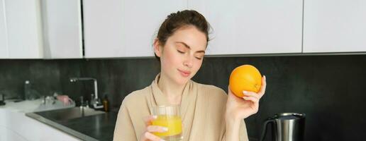 retrato de hermoso, sano joven mujer con naranja en mano, bebidas recién exprimido jugo desde vaso, posando en cocina, disfruta su Mañana a hogar foto