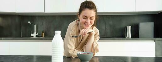 retrato de hermosa joven y sano mujer en bata de baño come su desayuno en cocina, tiene cereales con Leche y sonriente foto