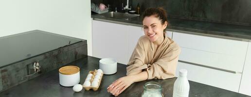 Attractive young cheerful girl baking at the kitchen, making dough, holding recipe book, having ideas photo