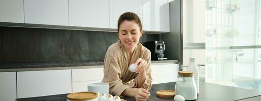 Attractive young cheerful girl baking at the kitchen, making dough, holding recipe book, having ideas photo