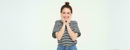 retrato de linda sonriente niña rubor, sostiene manos en las mejillas y mira entusiasmado, recibir contento noticias, soportes terminado blanco antecedentes foto