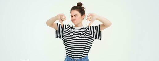 Portrait of disappointed, young modern woman, student shows thumbs down, dislikes smth, disapproves, stands over white background photo