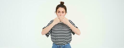 Image of shocked woman, covers her mouth, looks surprised, stands over white background photo