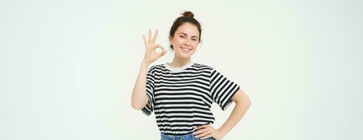 No problem, excellent choice. Smiling, confident young woman, showing okay, ok sign, zero gesture, recommends product, stands over white background photo