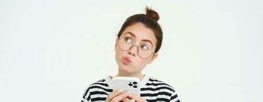 Online shopping, social media and cellular technology concept. Young woman in glasses, holding smartphone, thinking, using mobile phone, standing over white background photo
