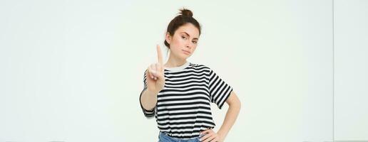 Image of confident woman, shows one finger, taboo, stop gesture, disapprove smth, gives warning, stands over white background photo