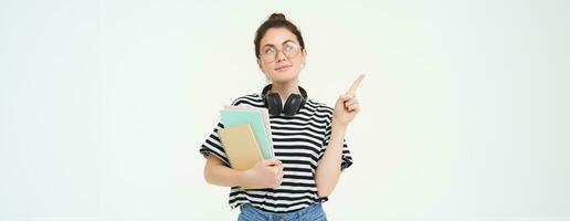 Image of smart young woman in glasses, student carries her notebooks and planners, points left with thinking face, making choice, deciding on smth, white background photo