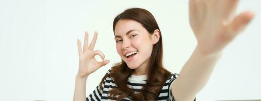 Portrait of positive, happy young woman shows okay sign and takes selfie, holds smartphone with extended hand, posing near something she recommends, isolated on white background photo