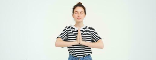 retrato de joven consciente de mujer meditando, sostiene manos abrochado juntos, namaste gesto, práctica yoga, en pie terminado blanco antecedentes foto