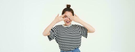 contento joven mujer, cumpleaños niña echar un vistazo mediante dedos, sostiene manos en ojos, esperando para sorpresa, soportes terminado blanco antecedentes foto