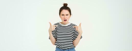Annoyed woman pointing at herself and frowning, arguing, looking frustrated and disappointed, shouting, standing over white background photo