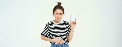 Woman takes painkillers, pills from stomach ache, menstrual pain, holds hand on belly, shows bottle with tablets, stands over white background photo