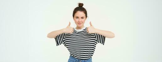 Portrait of confident, beautiful woman, showing thumbs up, recommends product, likes and approves, stands over white background photo