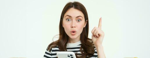Portrait of excited woman raising finger, eureka gesture, has an idea or solution, holds mobile phone, white background photo