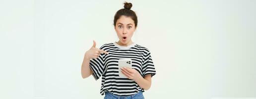 Portrait of impressed woman, drops jaw, points at her smartphone, sees amazing news online on mobile phone, isolated over white background photo