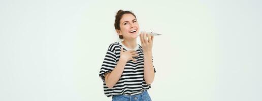 Happy young woman explaining something, using mobile phone app, talking on speakerphone, translating her voice, recording a message, holding telephone near lips, white background photo