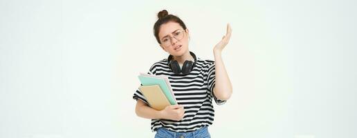 Portrait of compllicated, sad young woman in glasses, carries homework notebooks, facepalms, looks disappointed, stands over white background photo