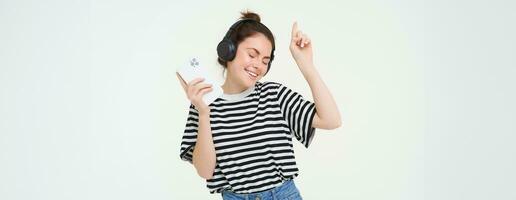 Portrait of happy woman with smartphone changes song on mobile phone streaming app, listens music in headphones, dancing against white background photo