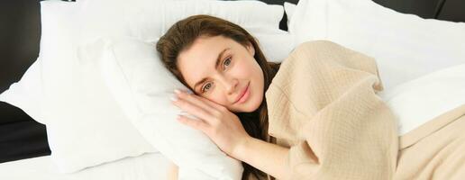 retrato de maravilloso mujer acostado en cama, descansando en almohada con ojos abrió, mirando a cámara y sonriente, vistiendo pijama, despertar arriba en Mañana foto