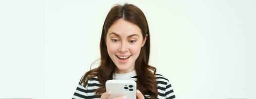 retrato de joven mujer mirando a teléfono inteligente pantalla con sorprendido, asombrado cara expresión, leyendo genial Noticias en teléfono foto