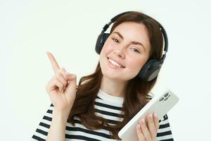 Portrait of smiling student, girl in headphones, holding mobile phone, pointing left, showing advertisement, store offer, white background photo