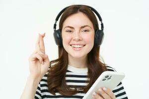 Portrait of smiling female model using headphones and smartphone, hold fingers crossed, makes wish, hopes for something, anticipates, stands over white background photo