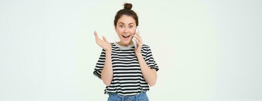 Portrait of girl answers phone call, reacts surprise, stands over white background. Communication and lifestyle concept photo