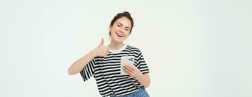 Enthusiastic girl with smartphone shows thumbs up, isolated over white background photo
