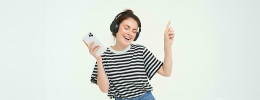 Young woman with smartphone listening to music, dancing to her favourite song in headphones, posing against white background photo