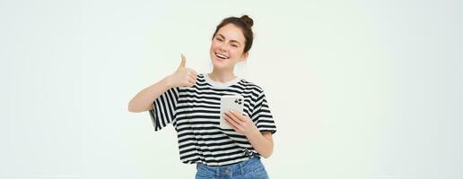 Smiling young woman showing thumbs up in approval, recommends app, holds smartphone, looks satisfied, isolated over white background photo