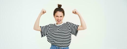 Image of sassy, strong and confident woman shows her muscles, flexing biceps, raising her arms high, standing over white background photo