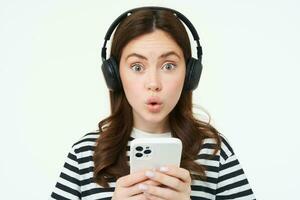 retrato de contento joven moderno mujer, hembra modelo en inalámbrico auriculares, sonriente y mirando a cámara, participación teléfono inteligente, utilizando móvil teléfono aplicación, blanco antecedentes foto