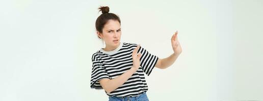 Image of woman expressing aversion, dislike something, looks apalled, extends hands forward in defensive gesture, rejecting, refusing, standing over white background photo