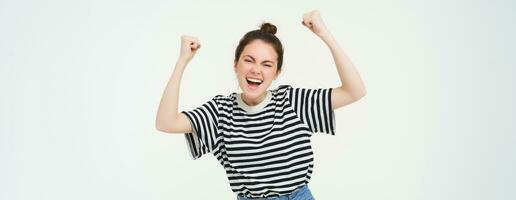 Portrait of happy young sports girl, fan rooting for team, celebrating victory, raising hands up, chanting, triumphing, standing over white background photo