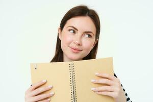 Portrait of beautiful young woman, holding daily planner, writing in her diary, making notes in notebook and smiling, dreaming about something, standing thoughtful against white background photo