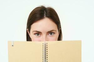 Portrait of cute young woman hides her face behind planner, holds notebook against her face and smiles, isolated over white background photo