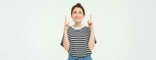 Portrait of excited, smiling young woman, looking and pointing up with happy, emotional face, white background photo