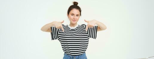 Portrait of confident, smiling brunette woman, pointing fingers down, showing banner on bottom, follow link below gesture, standing over white background photo